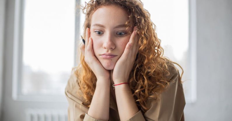 Task Lighting - Pensive female student with hands on cheeks looking down while sitting in light room on blurred background while deciding task in frustration